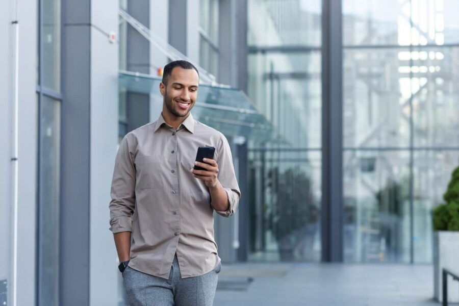 Smiling man in a smart-casual outfit walking outside a modern glass building while checking his smartphone
