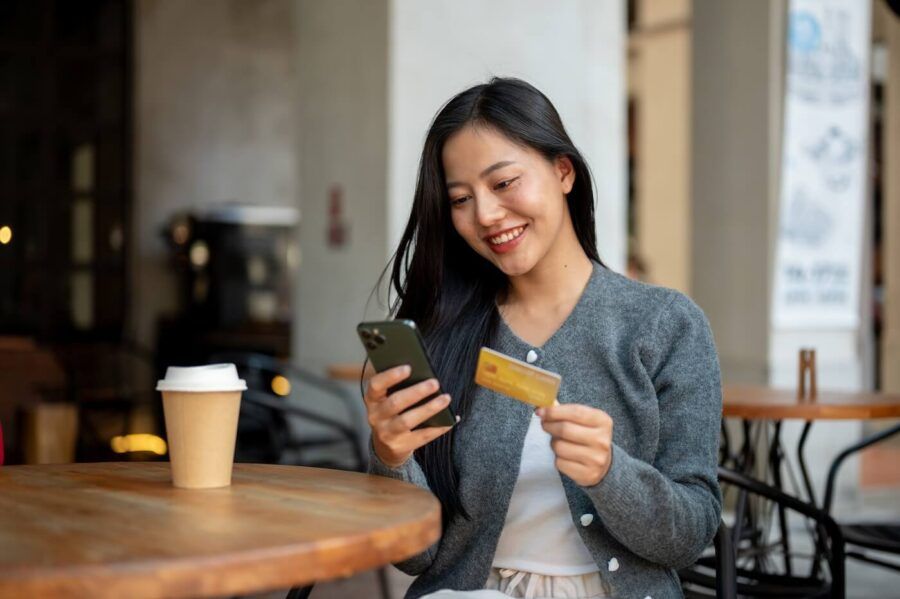 Smiling young woman enjoying coffee at a café while using her smartphone and holding a credit card