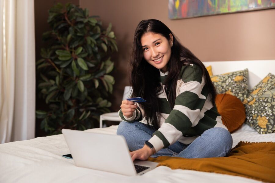 Smiling woman holding new credit card in hands sitting on bed in front of laptop with credit card.