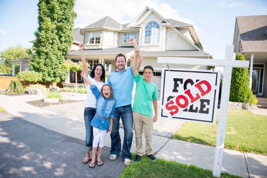 A family of four standing on a sidewalk in front of a house with a “For Sale” sign displaying a “Sold” label, with arms raised, and a single‑family home visible in the background.