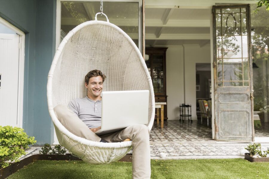 Happy young man using laptop on swing chair in his backyard.