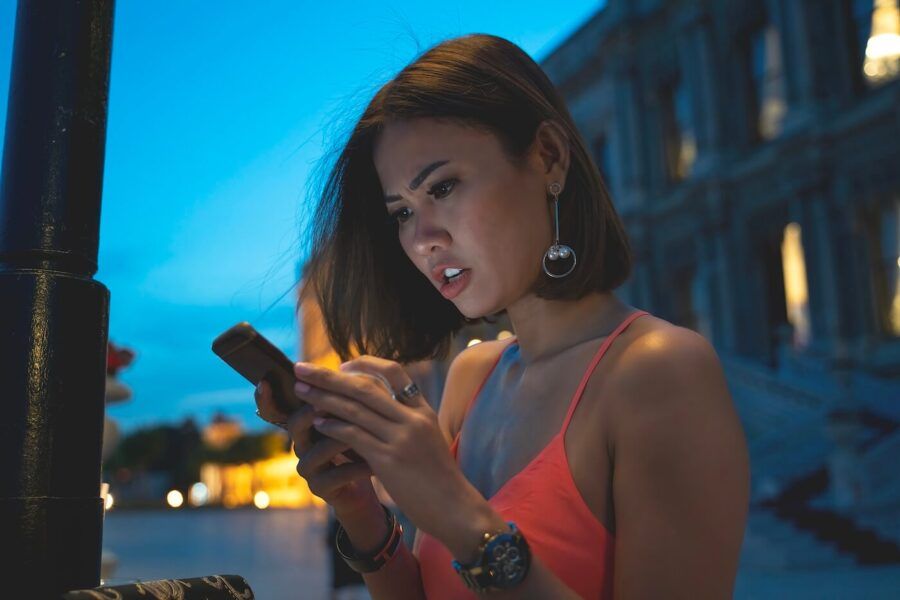 A concerned young woman standing on a dimly lit street at night, looking at her smartphone