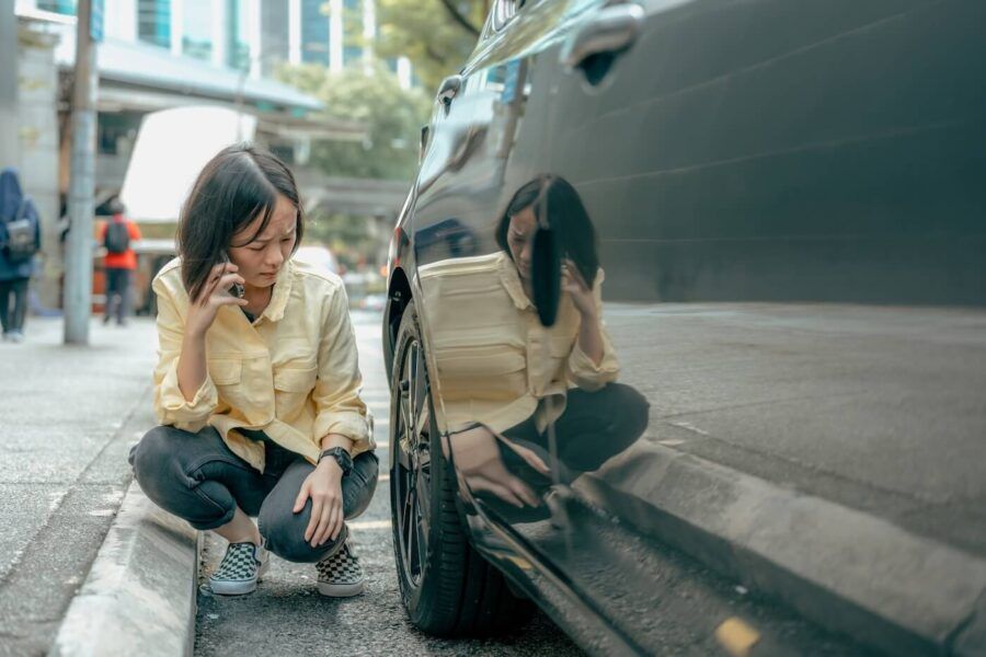 A concerned woman squatting next to her car, examining visible damage while speaking on the phone with her insurance company