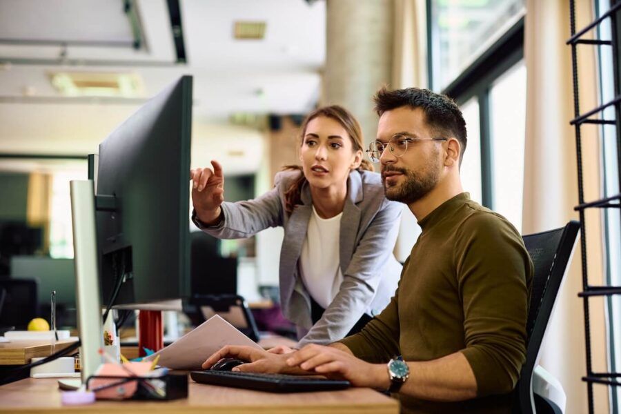 Two coworkers using a computer; the woman is pointing at a monitor while the man while uses a keyboard and holds documents.