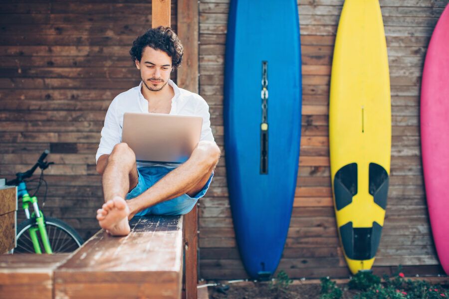 Young man using laptop outdoors in the summer with surf boards in the background.