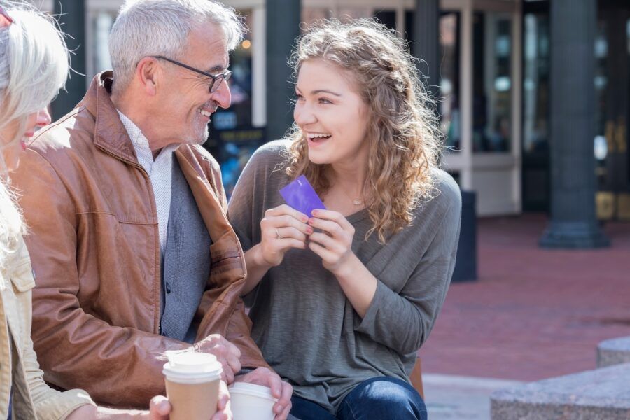 A young woman sits with her grandparents along a city street. She smiles gratefully as she holds up the debit card they have just given her.