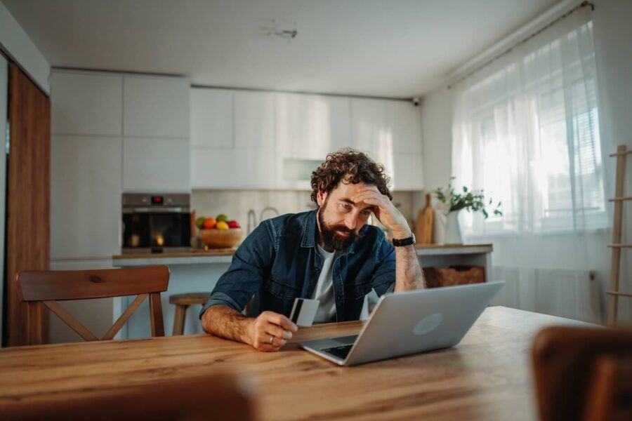 Concerned man sitting at the kitchen table, holding a credit card and looking at the laptop screen