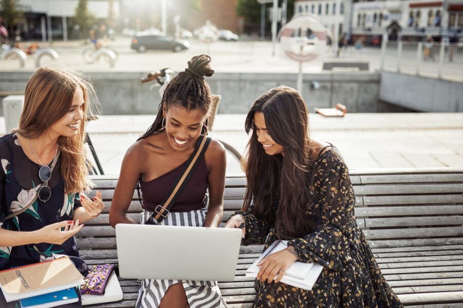 Shot of three young women looking at a laptop together outdoors on a park bench.