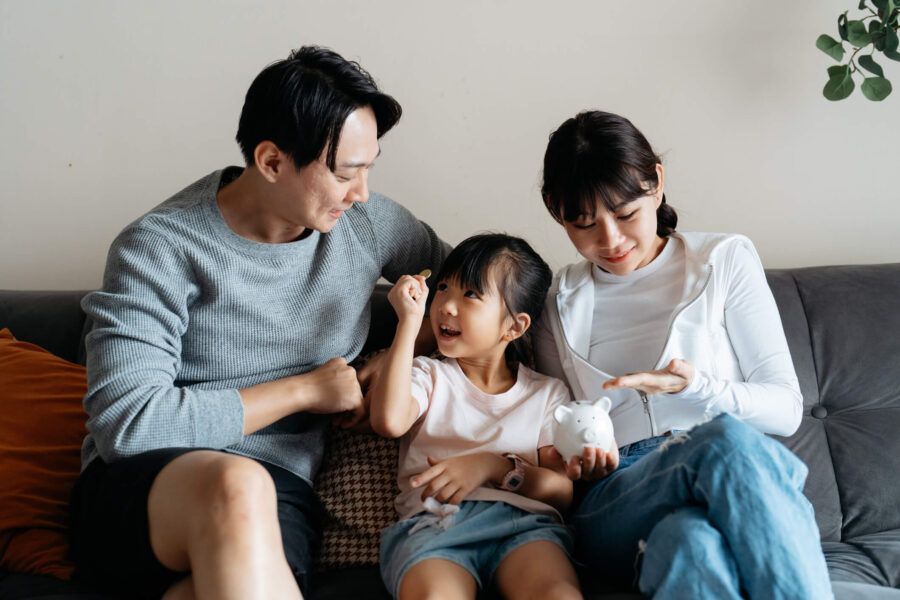 Happy family with a toddler girl sitting on the sofa; the girl is showing a coin to her father while the mother is holding a piggybank