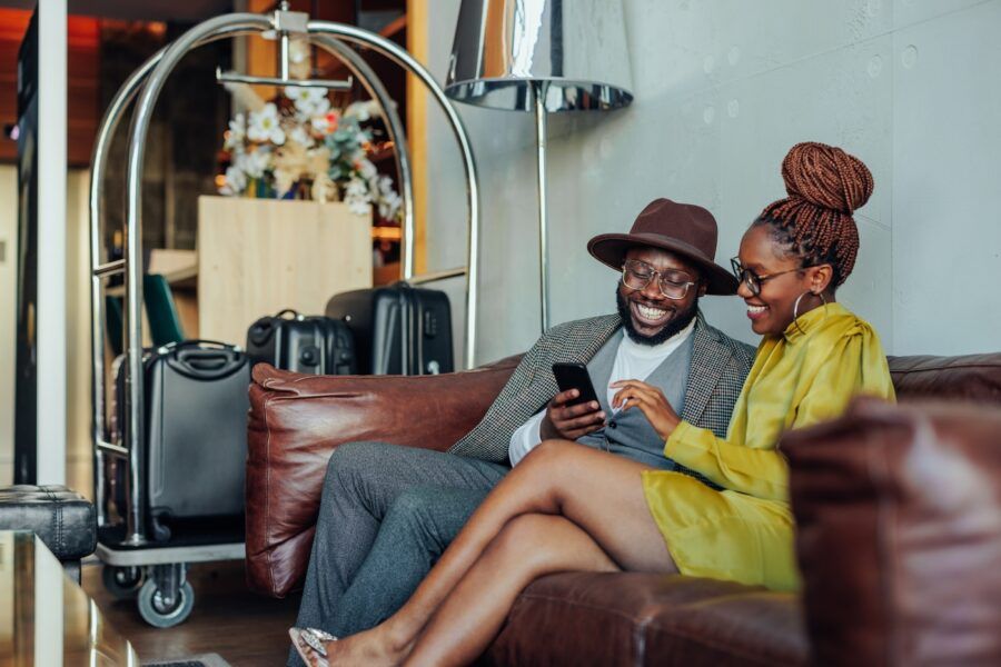 A wealthy couple is lounging in the hotel as they're waiting to be taken to their luxurious suite in the hotel. They are sitting on a sofa with their luggage on the cart and are using a smartphone.
