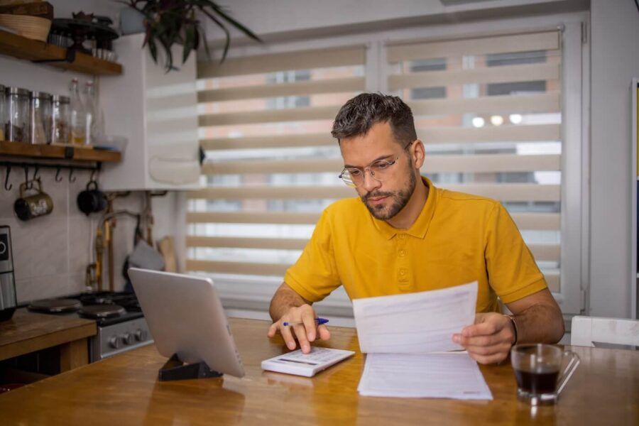 Focused man sitting at the dining table and using calculator while holding a printed document, with a tablet standing next to him