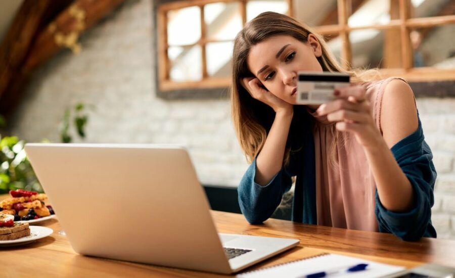 Pensive young woman looking at her credit card while using her laptop at home