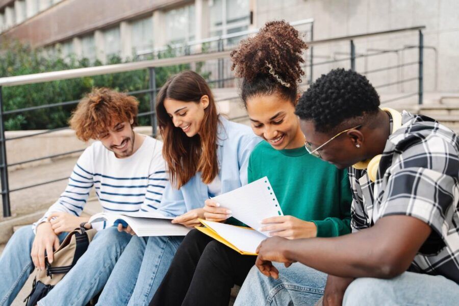 Group of two female and two male students sitting on campus stairs, studying with books and notepads