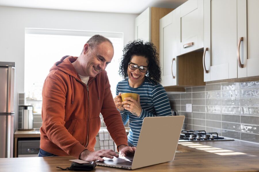 A shot of a happy couple standing in their kitchen at home. They are looking at their computer together and making plans for the future. The woman is holding a hot beverage in her hand.