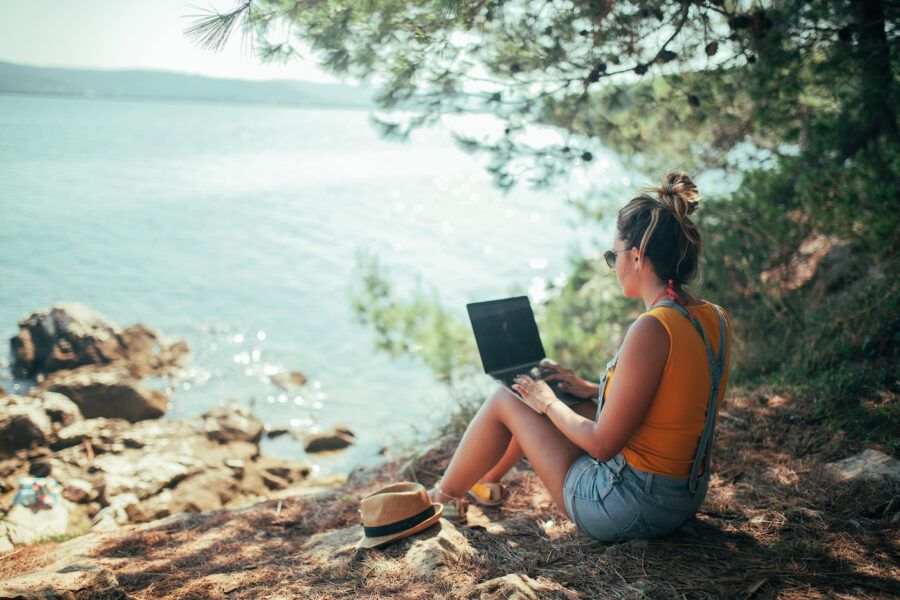 A woman sitting on the shore facing the ocean and holding a laptop.