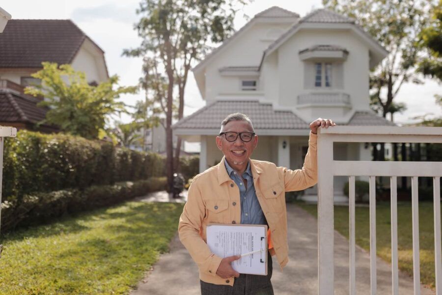 Smiling male appraiser leans against a fence while holding a notepad, standing in front of a charming old white house.