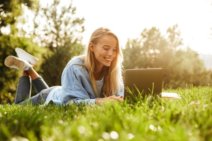 Portrait of a smiling young girl laying on a grass at the park, using laptop computer.