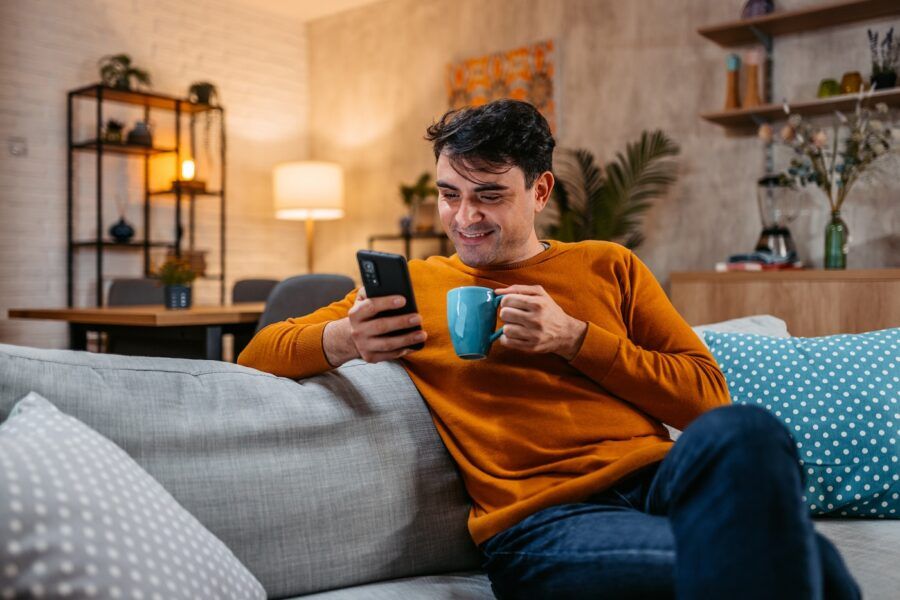 Smiling man relaxing at home, reading on smartphone and drinking coffee.
