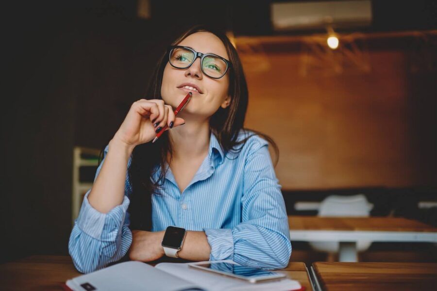 A smiling young woman thinking while holding the pen at her chin