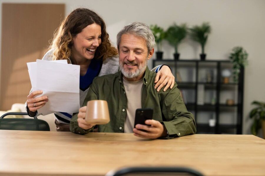 Mature couple sitting at a table, with the woman holding papers and the man holding a phone and a mug, in a room with shelves and plants in the background
