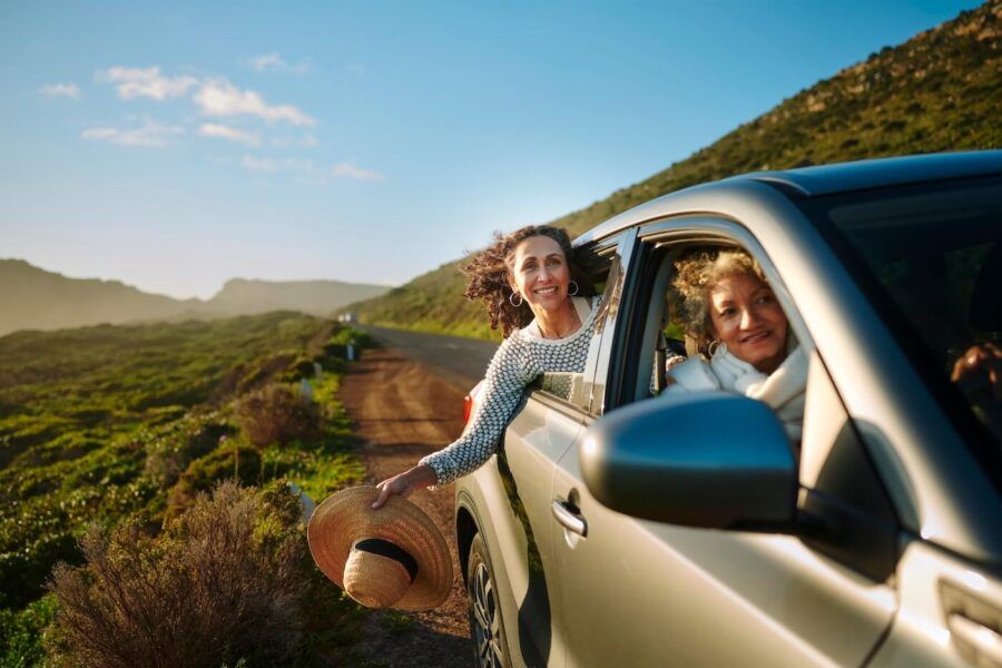 Two women riding in a car with the windows down, driving in the sunny countryside.