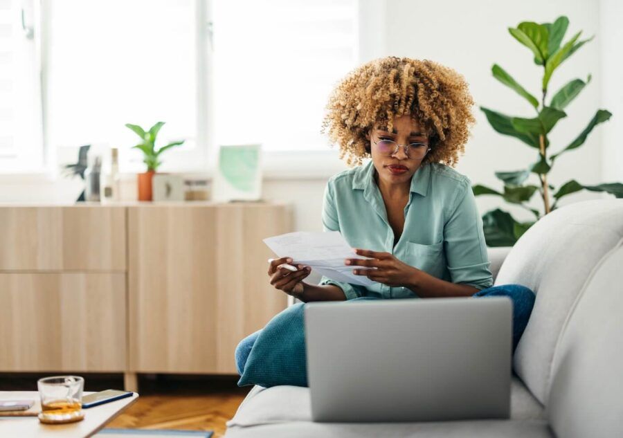 Focused young woman using her laptop to pay bills while seating on a couch in a bright living room