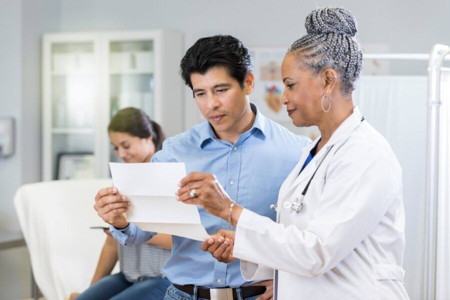 Smiling female doctor reviewing test results with a female patient’s partner, while the patient sits in the background