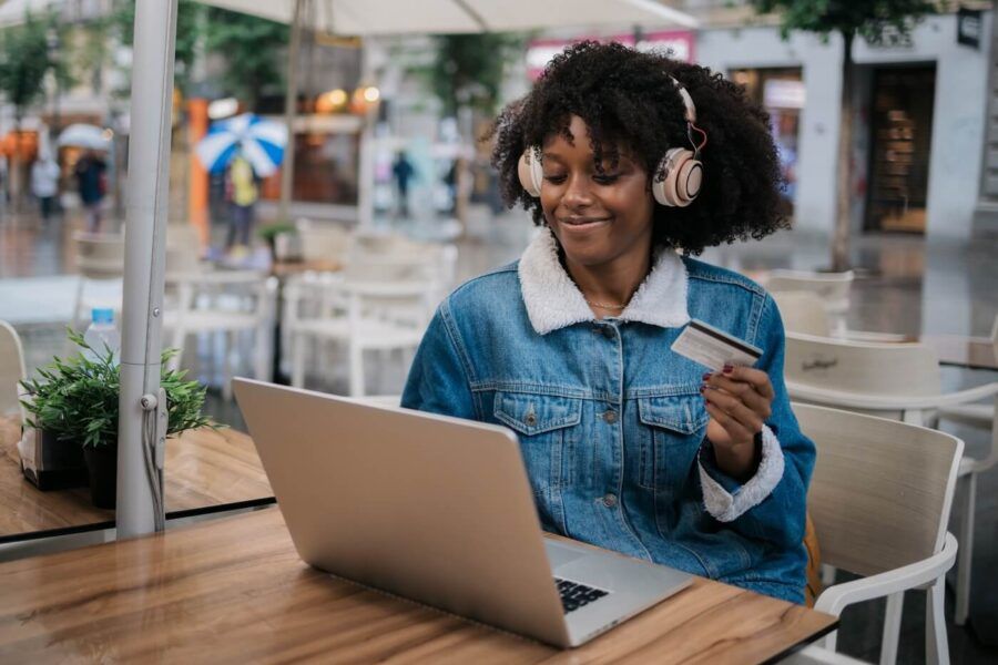 Smiling young woman wearing headphones at an outdoor café, using her laptop to make an online purchase with a credit card
