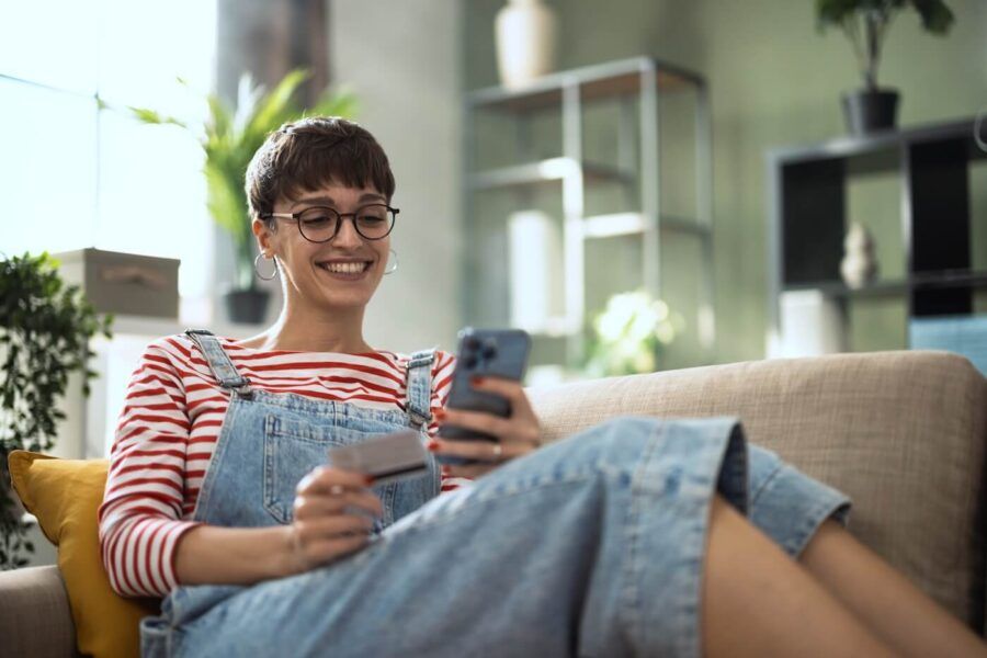 Smiling woman sitting comfortably on the sofa while holding her credit card and the smartphone