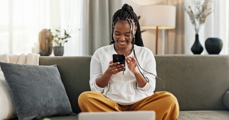 A smiling woman looking at her investing accounts on her phone while sitting on the couch.