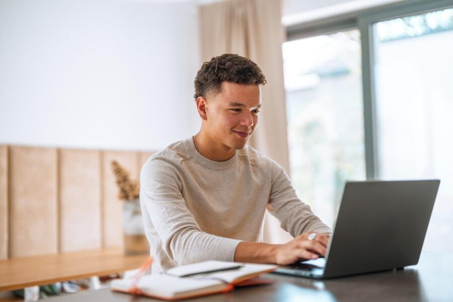 Smiling young man works on a laptop at a table in a bright room, with a notebook and pen nearby.
