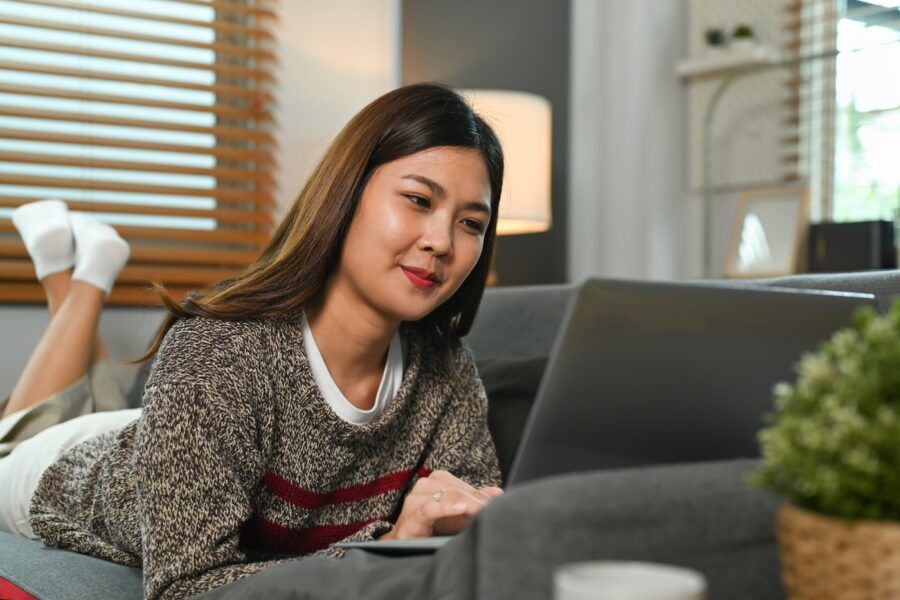 Smiling woman wearing warm sweater using laptop on comfortable couch at home.
