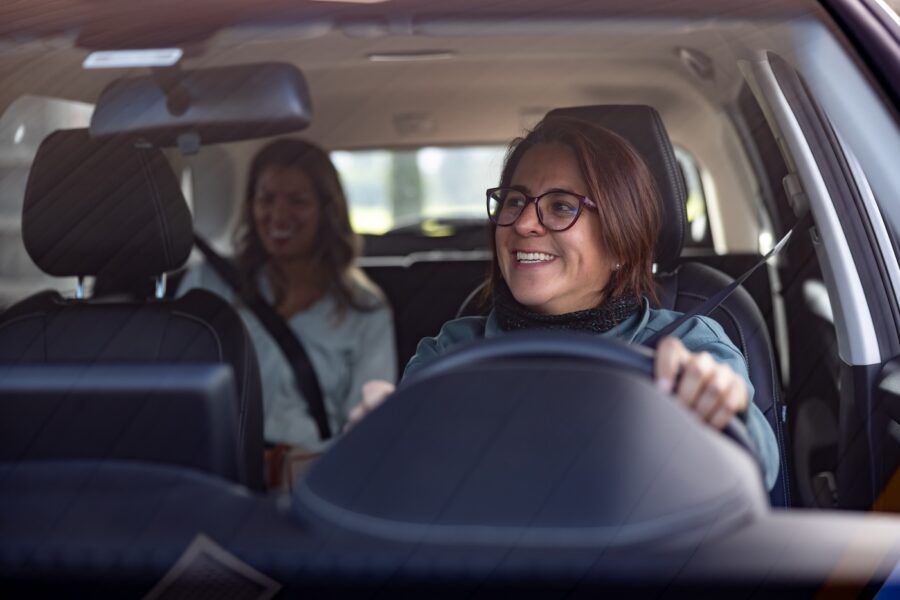 Happy female crowdsourced taxi driver talking to a female passenger in the back seat and looking through the rear view mirror
