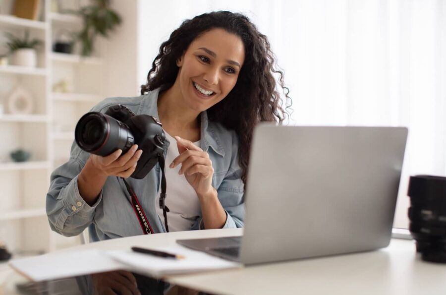 Smiling woman holding a photo camera while using her laptop in a home office