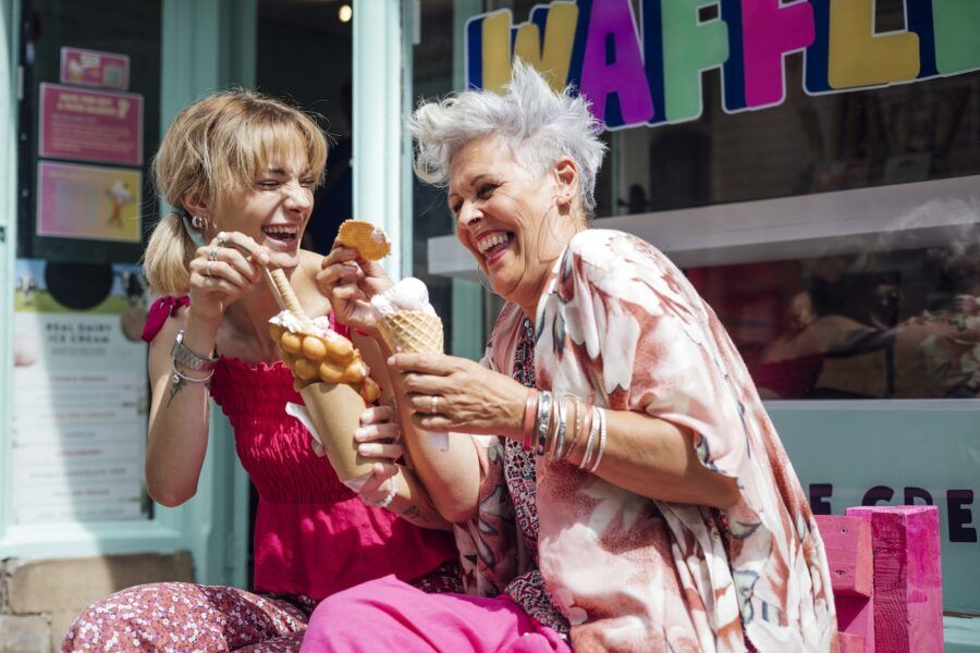 Two women sitting side by side on a bench outside of a cafe store eating ice cream and waffles together. They are enjoying the sunshine, laughing and talking together.