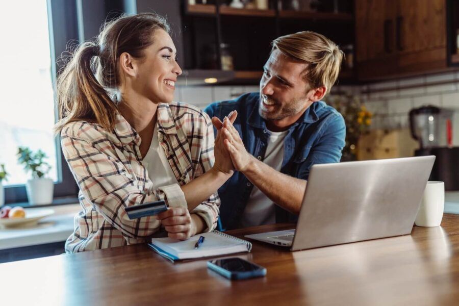 Happy couple sits in the kitchen with an open laptop, giving a high five; the woman holds a credit card