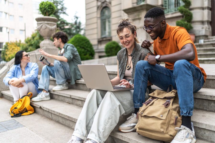 A group of college students sitting on outdoor steps using a laptop together, with backpacks nearby and a building in the background.