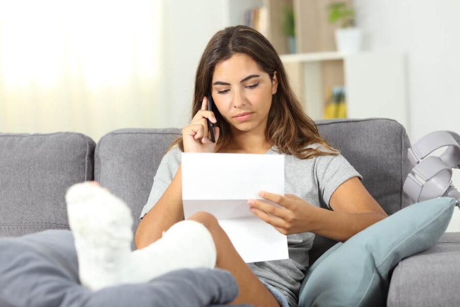Young woman with her leg in a cast elevated on a cushion, speaking on the phone while holding a medical bill