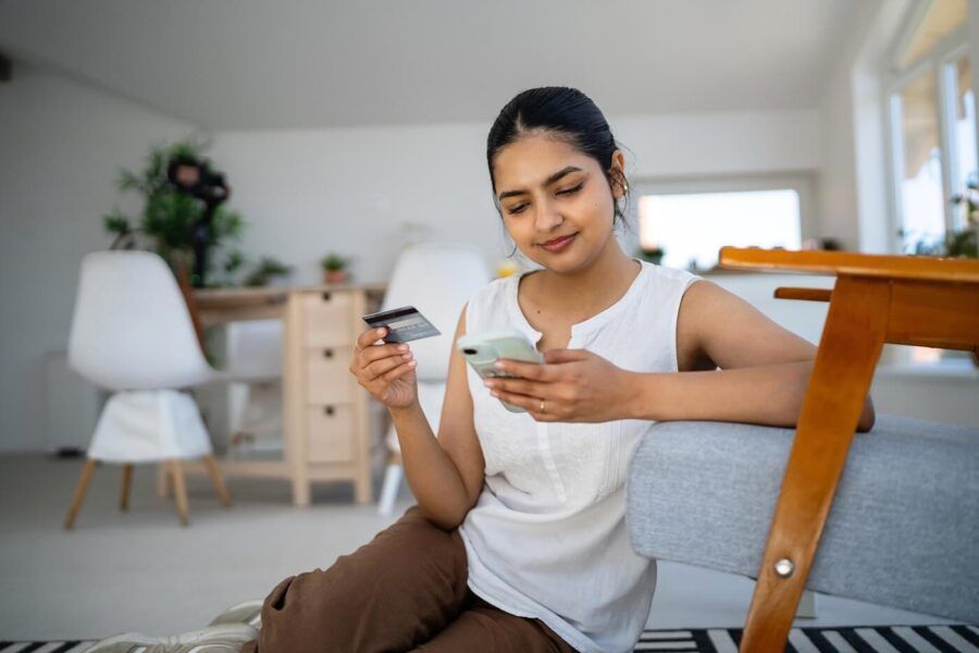 Confident young woman sitting on the floor in her living room and holding a debit card while checking her smartphone