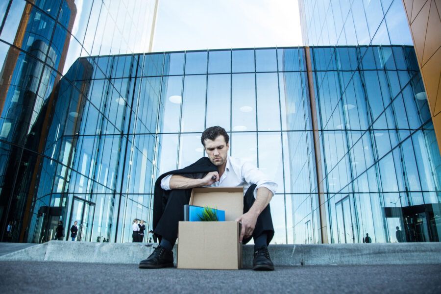 Fired business man sitting upset on the street near office building with a box of his belongings.