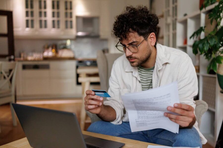 Focused man looking at his credit card while holding a printed document, with an open laptop beside him on a coffee table