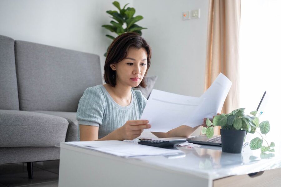 Focused woman reviews paperwork while seated at a coffee table, with a laptop and calculator nearby, in a bright living room with a sofa and houseplants in the background