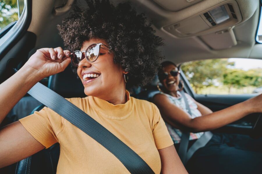 Happy woman in sunglasses sitting on a passenger seat and looking in the car window; another smiling woman is driving a car