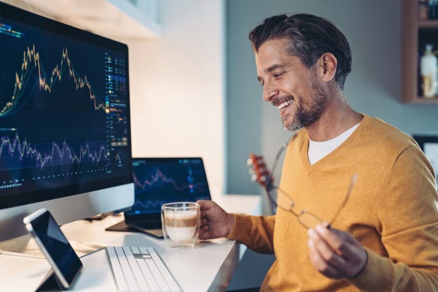 A smiling man is reviewing the cryptocurrency prices on the monitor while having a coffee