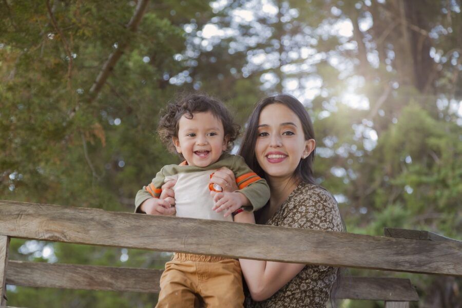 A young woman with her smiling baby, lifting him up next to a wooden gate in a park.