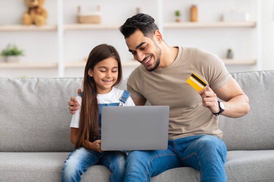 Portrait of smiling family doing online shopping at home using laptop, father holding credit card in hand, dad and daughter sitting on couch at home