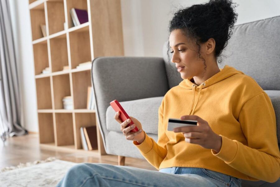 Focused young woman sitting on the floor in her living room and using her smartphone and credit card