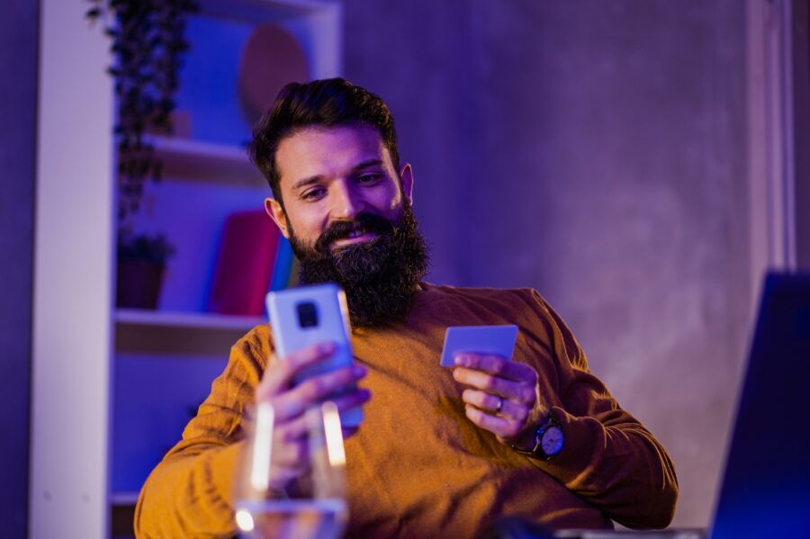 A cheerful man is sitting at a blue and purple lighted home office and paying online on his phone while smiling at it.