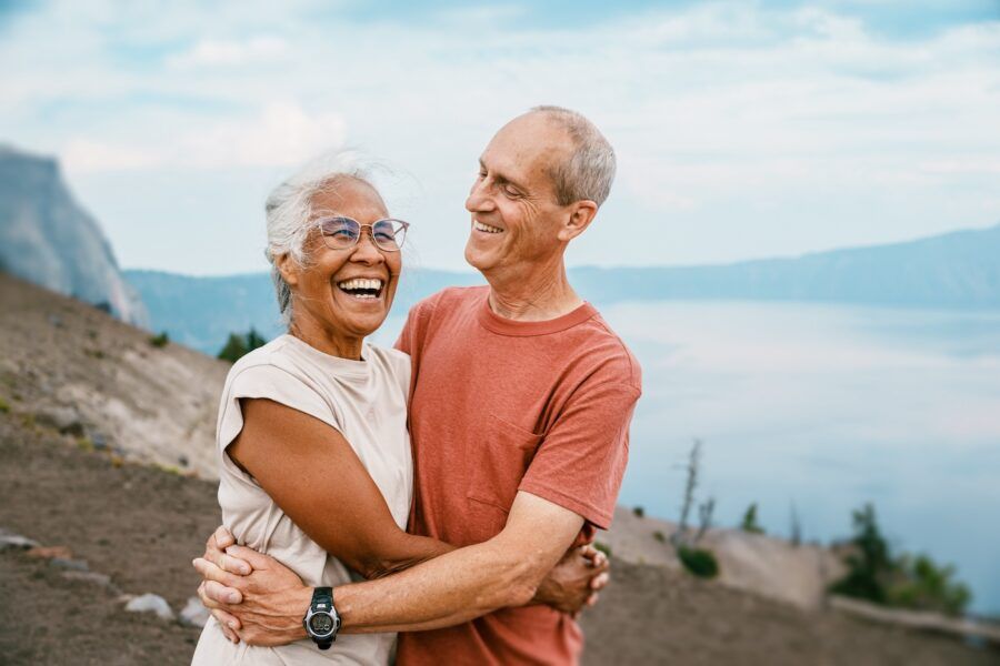 A vibrant senior woman laughs and affectionately embraces her husband while out on a mountain hike overlooking a lake.
