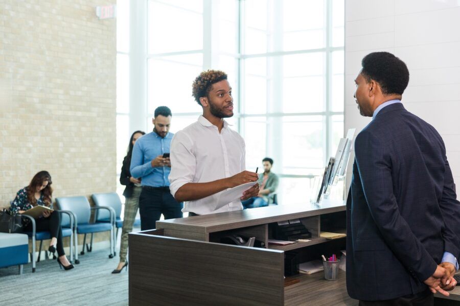 A young adult man holding paperwork listens as the mature adult male bank teller explains how to withdraw money from savings.